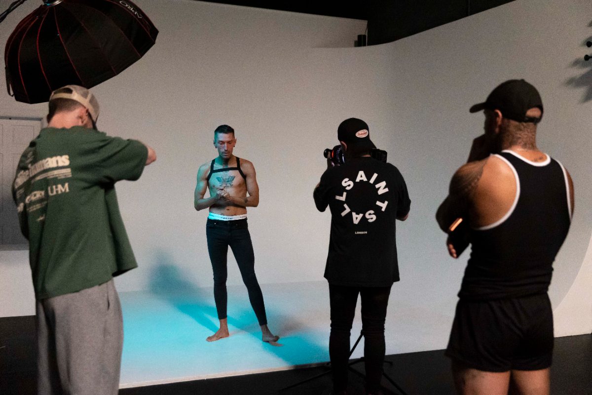A client stands against a black studio backdrop while the photographer and lighting rig frame the shot